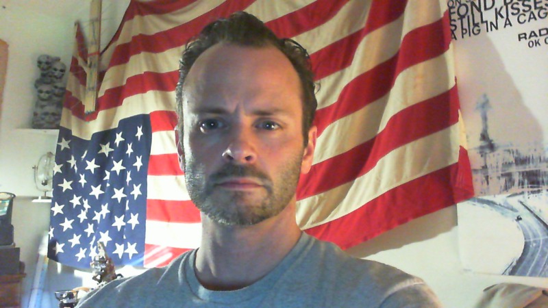 Bust photo of dark-haired blue-eyed middle-aged man afront an upside-down American flag